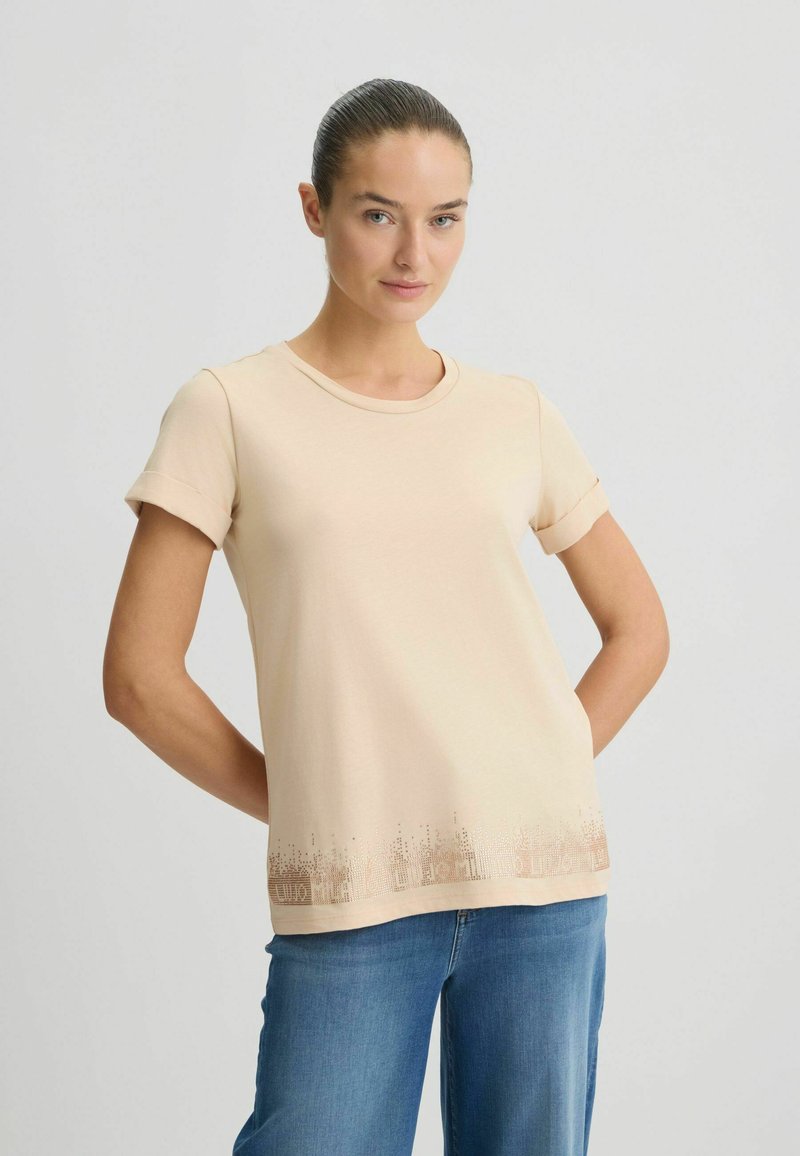 Beige t-shirt met korte mouwen en een ronde halslijn, voorzien van een subtiel gemêleerd patroon langs de rand. Gecombineerd met spijkerbroeken.