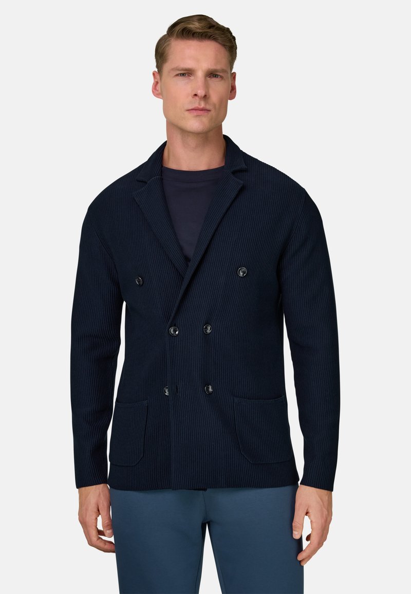 Marineblaue, zweireihige Jacke mit geripptem Muster, zwei vorderen Taschen und schwarzen Knöpfen. Kombiniert mit einem dunklen Hemd und hellblauen Hosen.