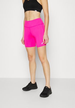 Mujer con pantalones cortos deportivos de color rosa brillante, sujetador deportivo negro y zapatillas negras, de pie frente a un fondo liso y claro.