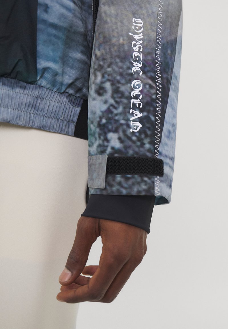 Windbreaker mit einem blauen und grauen abstrakten Muster, aus strukturiertem Stoff, mit eng anliegenden Bündchen und einem Klettverschlussband am Ärmel.