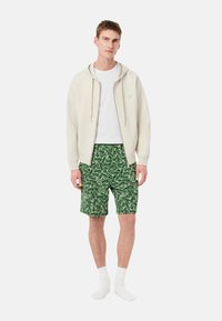 Hoodie beige chiaro con cordini, t-shirt bianca sotto, pantaloni corti verdi con motivo a foglia, abbinati a calze bianche.