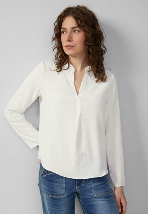 Weißes Langarmshirt mit V-Ausschnitt, weichem Stoff und leicht geschwungenem Saum, kombiniert mit blauen Jeans.