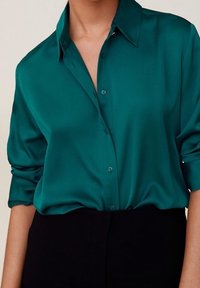 Teal green silk long-sleeve button-up shirt with collar, partially tucked into black high-waist pants, worn by a person with rolled sleeves.