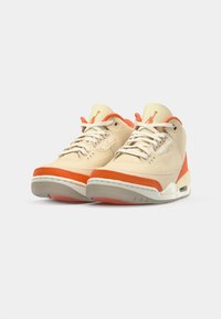 Zapatillas beige con acentos naranjas, que cuentan con una parte superior de tela texturizada, cordones blancos y una suela de goma gris con amortiguación de aire visible.
