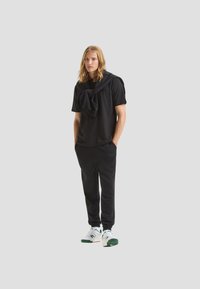 Outfit con t-shirt nera e pantaloni da jogging, caratterizzato da un maglione nero legato sulle spalle, abbinato a scarpe da ginnastica bianche e verdi.
