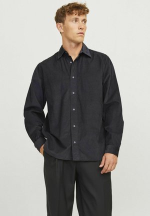Camisa oscura de botones, hecha de tela texturizada con un ajuste relajado, que presenta un cuello clásico y botones morados, combinada con pantalones negros.