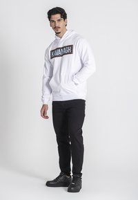 Sudadera blanca con un diseño de "KAVANAGH" en letras marrones y azules, combinada con jeans negros y zapatillas negras. Textura de tela suave.