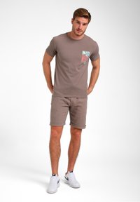 Korte mouwen bruine t-shirt met een kleurrijk patroon op de pocket, gecombineerd met bijpassende bruine shorts en witte sneakers.