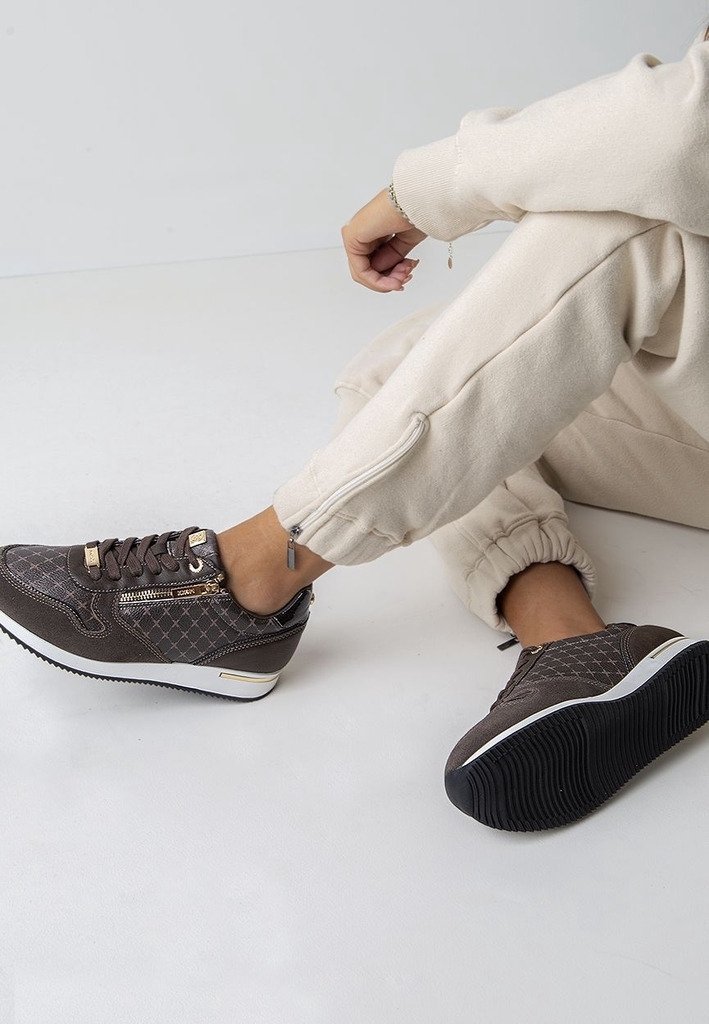 Personne en jogging beige assise par terre portant des baskets marron foncé à motifs avec fermeture éclair dorée et semelles blanches.