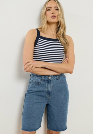 Lyshåret kvinde med løst hår iført ærmeløs navy- og hvidstribet top og højtaljede denimshorts med rå kanter, armene krydset.