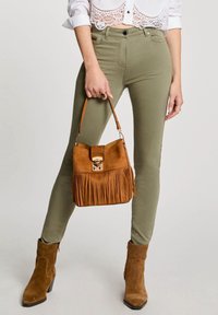 Olivově zelené skinny kalhoty s vysokým pasem, doplněné hnědou surovou kabelkou s třásněmi a zlatým zapínáním, a hnědými koženými kotníkovými botami na podpatku.