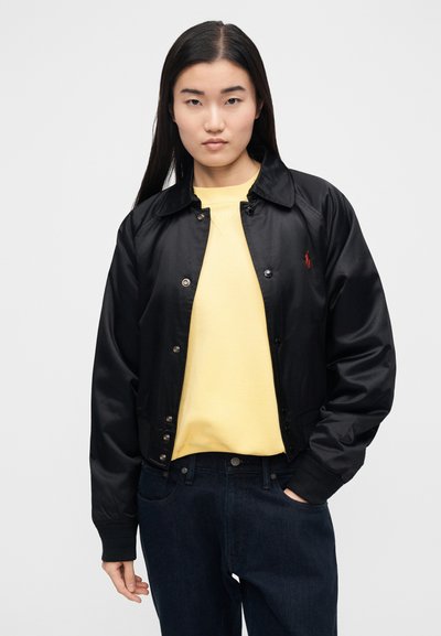 Čierna bombera bunda so zapínaním na stláčacie gombíky, golierom a rebrovanými manžetami. Nosená cez žltú mikinu a v kombinácii s tmavými džínsami.