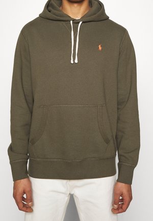 Sweat à capuche vert olive avec cordons blancs, poche kangourou et petit logo orange sur la poitrine gauche, porté avec un pantalon blanc.