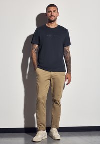 Muž s tetovanými pažemi stojí proti bílé zdi, oblečený v námořnickém modrém tričku, béžových kalhotách a bílých teniskách, s rukama v kapsách.