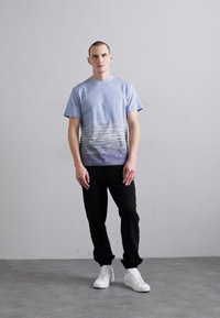 Blå gradient t-skjorte med falmende horisontale linjer, korte ermer, kombinert med svarte bukser og hvite sneakers, stående mot en grå bakgrunn.
