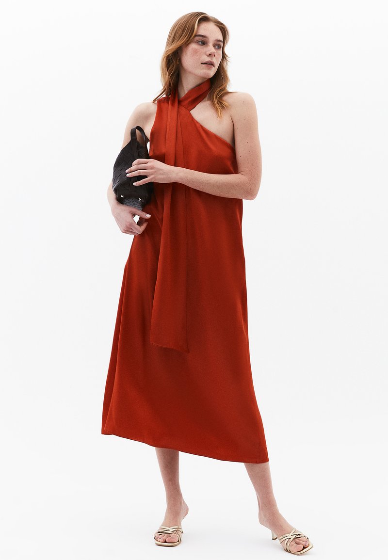 Rotes Halfterkleid aus glattem Stoff, Midi-Länge, mit fließender Silhouette. Das Model hält eine strukturierte schwarze Tasche und trägt leichte Sandalen.