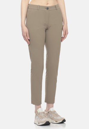 Pantaloni beige slim fit corti, indossati con sneakers beige e bianche su una persona in piedi contro uno sfondo semplice.