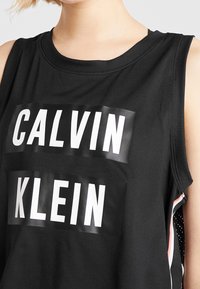 Mustat hihattomat urheilupaidat, joissa on suuret valkoiset "CALVIN" ja "KLEIN" -logot. Sujuva kangas, jossa on pyöreä kaula-aukko ja raidalliset sivuelementit.