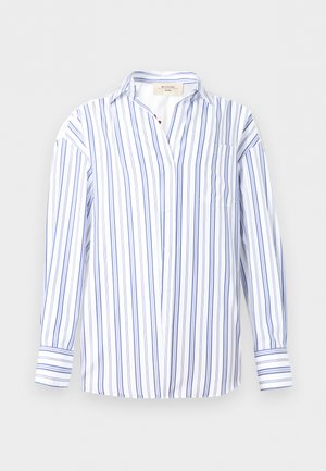 Γυναίκα που φοράει μπλε και λευκό ριγέ μπλουζάκι μακρυμάνικο, μπεζ σορτς και μαύρα σαγιονάρες, στέκεται με το ένα χέρι στην τσέπη.