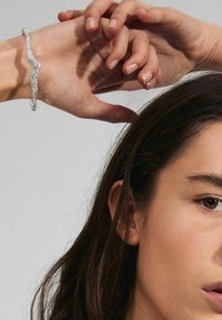 Hopeaketju hopeasta, jossa on aaltoileva muotoilu ja teksturoitu pinta, koristaa kättä, jossa on lyhyet, luonnolliset kynnet. Taustana on neutraali harmaa.