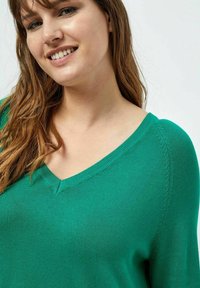 Maglione verde a maglia con scollatura a V e dettagli di cuciture testurizzate. Il tessuto sembra morbido e aderente, evidenziando una silhouette fluida.