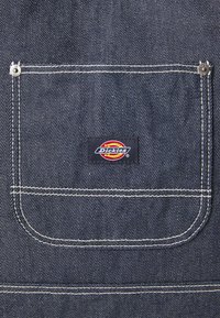Mörk denimtyg med en stor ficka med vit söm och en rektangulär svart Dickies-logotyp med röda och gula detaljer.