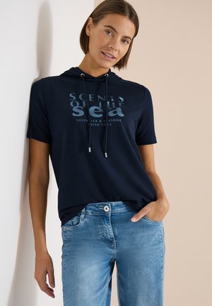 Vrouw draagt een marineblauwe korte mouwen hoodie met de tekst "Scents of the Sea", gecombineerd met lichtblauwe spijkerbroek, staand tegen een muur.