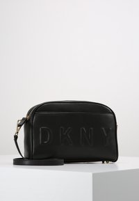Borsa a tracolla rettangolare nera con logo DKNY impresso, zipper dorato e tracolla regolabile, posizionata su una superficie bianca.