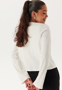 Top bianco a maniche lunghe con design cropped e maniche svasate. Il modello mostra la vista posteriore, con uno styling che presenta un materiale testurizzato.