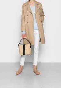 Beige Trenchcoat über einem hellblauen Pullover, weiße Hose, trägt eine Strohtasche mit schwarzen Akzenten. Sandalen vervollständigen das Outfit.