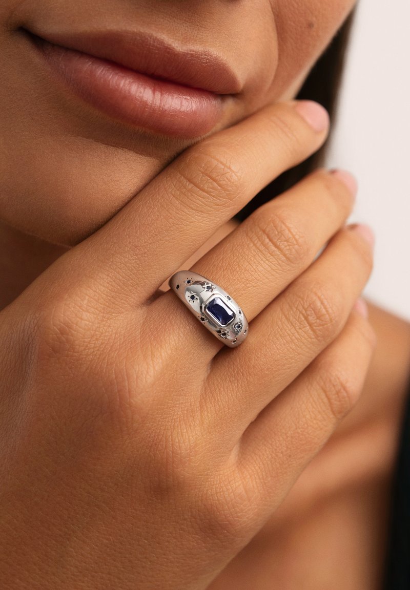 Primer plano de una mano tocando el mentón, que lleva un anillo de plata con una piedra rectangular de color azul oscuro y pequeñas grabaciones de estrellas.