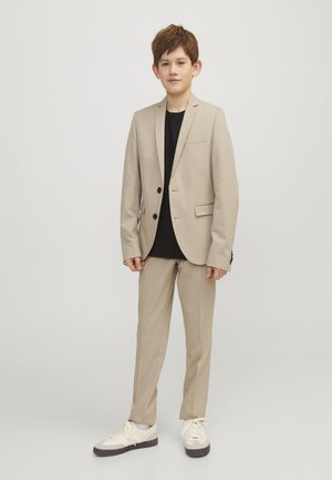 Boy standing in beige suit with black shirt and white sneakers against plain light background.