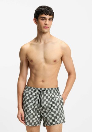 Giovane uomo che indossa pantaloncini da bagno a quadri bianchi e neri con la scritta "HUGO", in piedi con una mano in tasca contro uno sfondo bianco.