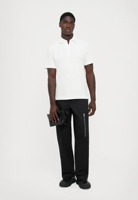 Hvit kortermet polo skjorte, svarte vide bukser med glidelåslomme, holder en svart clutch. Minimalistisk design med en jevn tekstur.