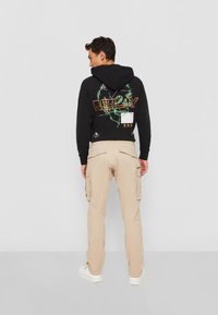 Sort hættejakke med grafisk design på ryggen, parret med beige cargo-bukser og hvide sneakers. Hættejakken har justerbar snoretræk.