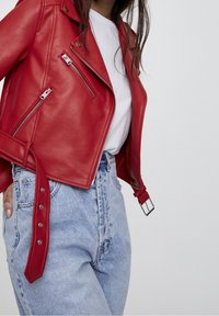 Femme portant une veste de motard en cuir rouge avec des fermetures éclair argentées sur une chemise blanche et un jean bleu clair taille haute sur un fond uni.