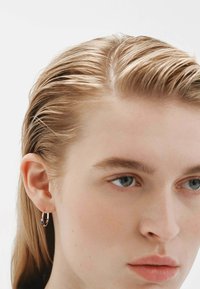 Pendiente de aro dorado con un acento de gema azul, cerca de la oreja de una persona con cabello lacio, castaño claro y piel clara.