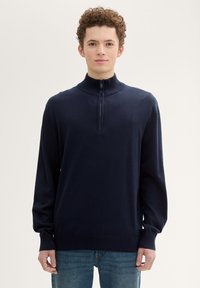 Pulover cu fermoar de culoare albastru marin, cu guler înalt, manșete din tricot și o textură netedă, asortat cu blugi de culoare albastră deschisă.