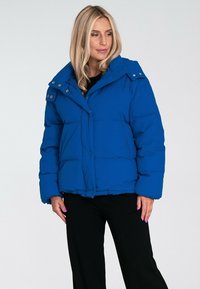 Niebieska kurtka puchowa z wysokim kołnierzem, detalami w postaci guzików oraz pikowanym wzorem. Posiada luźny krój i kieszenie z przodu.