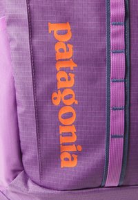 Paarse rugzak met een gestructureerde oppervlakte, oranje "patagonia" logo, en contrasterende roze en grijze banden.