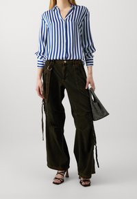 Camicia a righe blu e bianca con bottoni, pantaloni cargo larghi verdi scuri con tasche e lacci, abbinati a una piccola borsa nera in rete e tacchi neri.