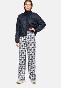 Marineblau gesteppte Jacke mit Knopfleiste, kombiniert mit weißen gemusterten Hosen mit Kettenmuster und schwarzen Akzenten.