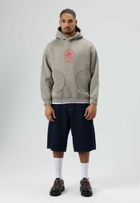 Sweat à capuche gris clair avec poches avant et logo rouge brodé. Associé à un short en denim foncé, des chaussettes blanches et des chaussures noires.