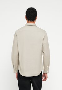 Camicia beige a maniche lunghe con colletto alla coreana con bottoni, texture liscia e cuciture ben visibili, indossata con pantaloni scuri.