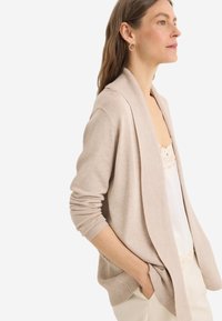 Cardigan beige con collo a scialle, polsini a coste e tasche, abbinato a un top in pizzo bianco, posizionato su uno sfondo neutro.