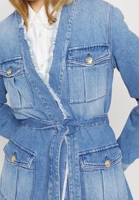 Abrigo de denim en azul claro con bordes deshilachados, dos bolsillos frontales con botones metálicos y un cinturón atado a la cintura. Camisa blanca abotonada debajo.
