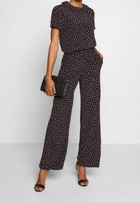 Combinaison noire avec col, manches courtes et petit imprimé cœurs rouges et blancs. Associée à une pochette noire et des sandales à talons.