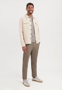 Licht beige, gestructureerde button-up jas met twee borstzakken, gecombineerd met een grijze geribde trui, tan kleurige broek en grijze sneakers met strepen.