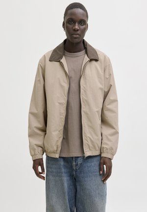 Fiatal személy bézs cipzáras dzsekit visel sötét gallérral, taupe színű pólót és bő, kék farmert, egy egyszínű fehér háttér előtt áll.