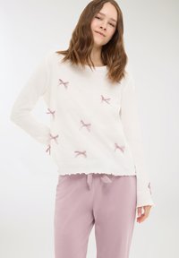 Giovane donna che indossa una maglietta bianca a maniche lunghe con piccoli fiocchi rosa abbinata a pantaloni viola chiaro con cordoncino su uno sfondo neutro.